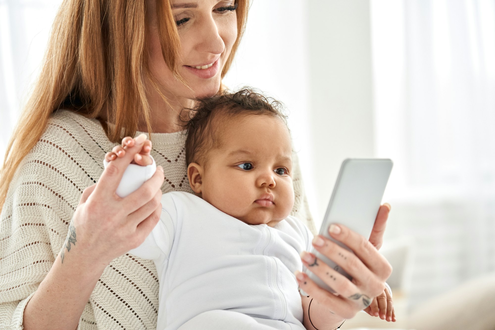 Happy young caucasian mother holding cute african baby daughter using phone.