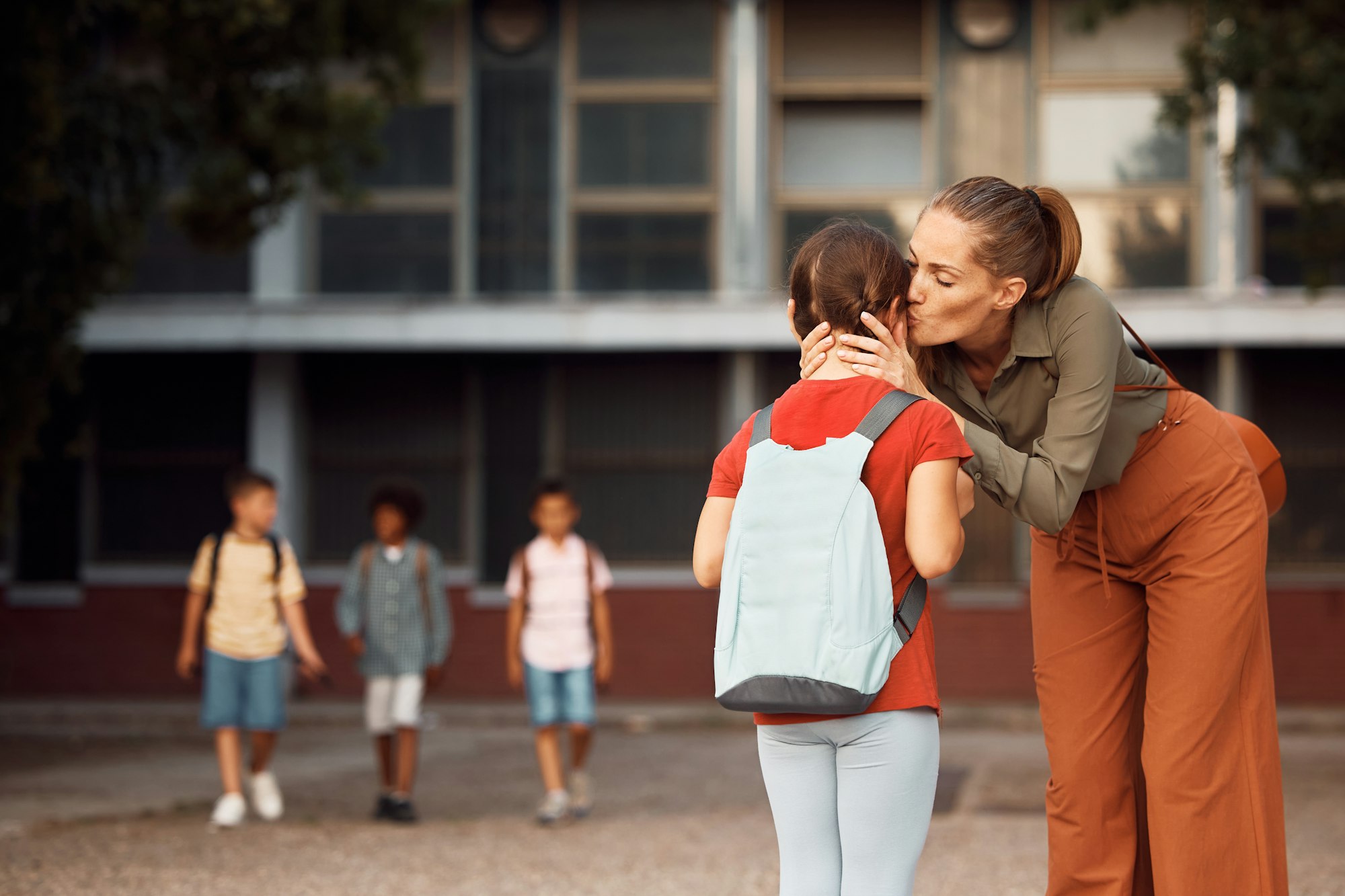 Loving mother kissing her daughter while greeting with her on first day of school in the schoolyard.
